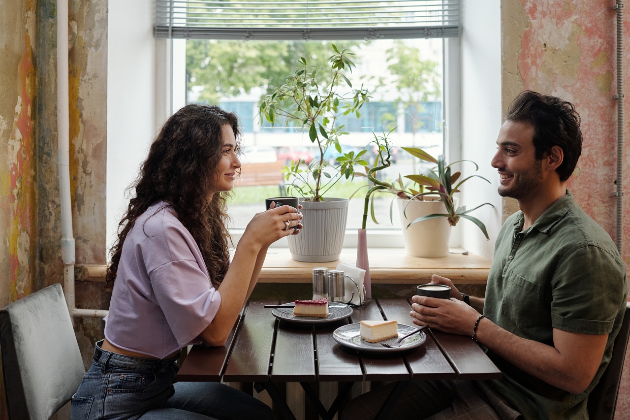 Young woman and her smiling boyfriend with cups of coffee sitting by table in front of one another and chatting