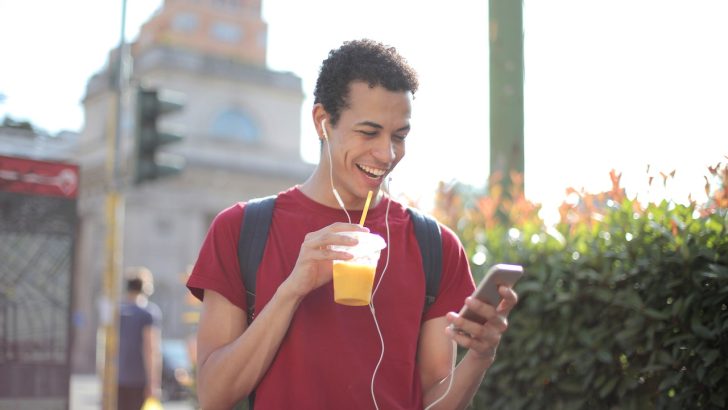 Cheerful man with smartphone drinking juice on street