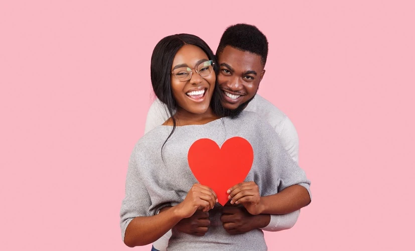 Black man and woman hugging and holding a red heart sign with a pastel pink background