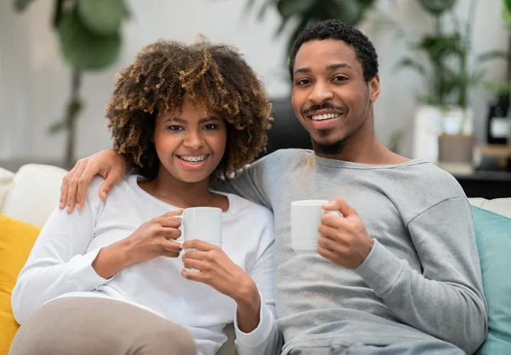 Black man and woman sitting down on a sofa hugging and holding a cup of coffee each