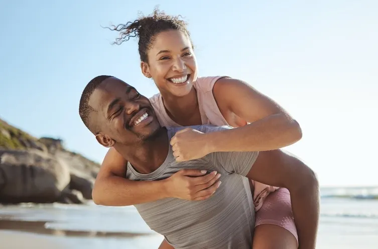 Black woman getting a piggy back on a black man while both smiling on a sunny beach with the sea and cliffs in the background
