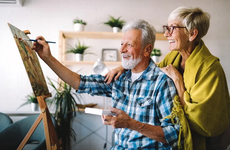 Older man and woman together smiling as they paint on a canvas in a sunlit room