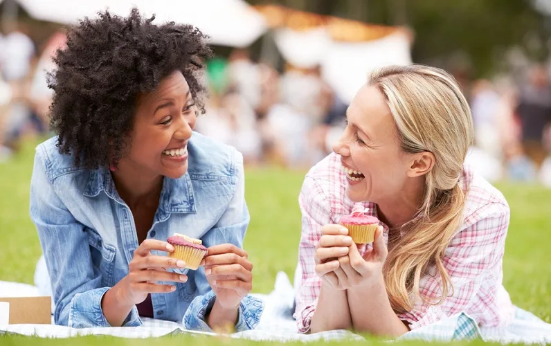 Two women laying down on picnic blankets looking at each other laughing and holding cupcakes
