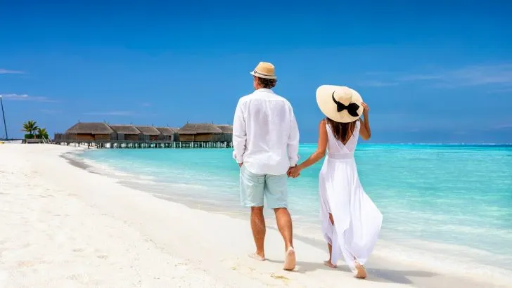 Happy couple in white clothing and with hats walks down a tropical beach with turquoise sea in the Maldives islands