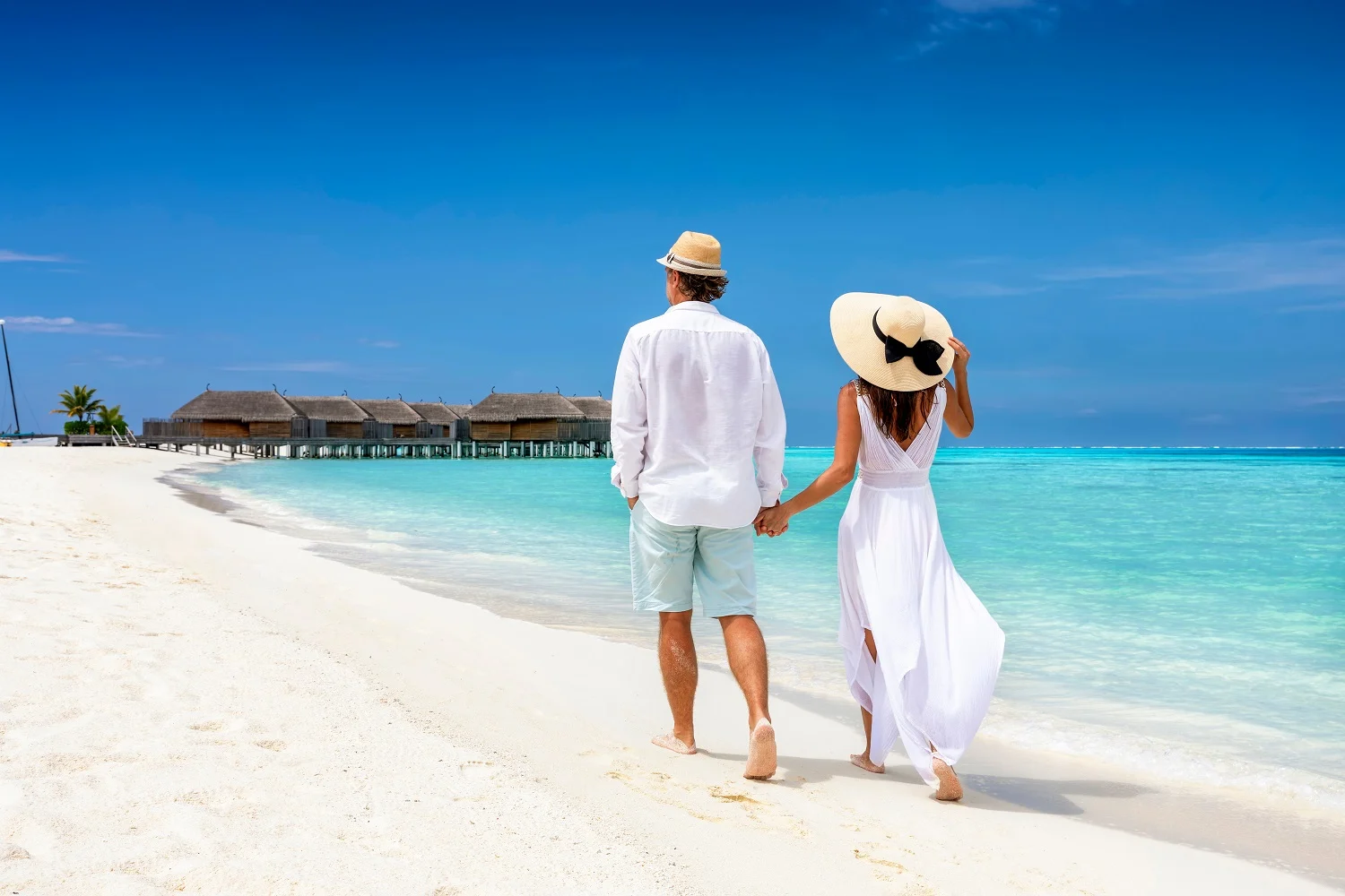 Happy couple in white clothing and with hats walks down a tropical beach with turquoise sea in the Maldives islands