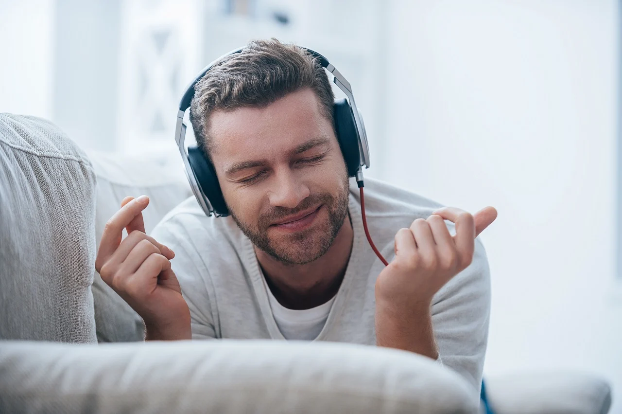 Cheerful young man in headphones listening to the music and gesturing while lying on his couch at home